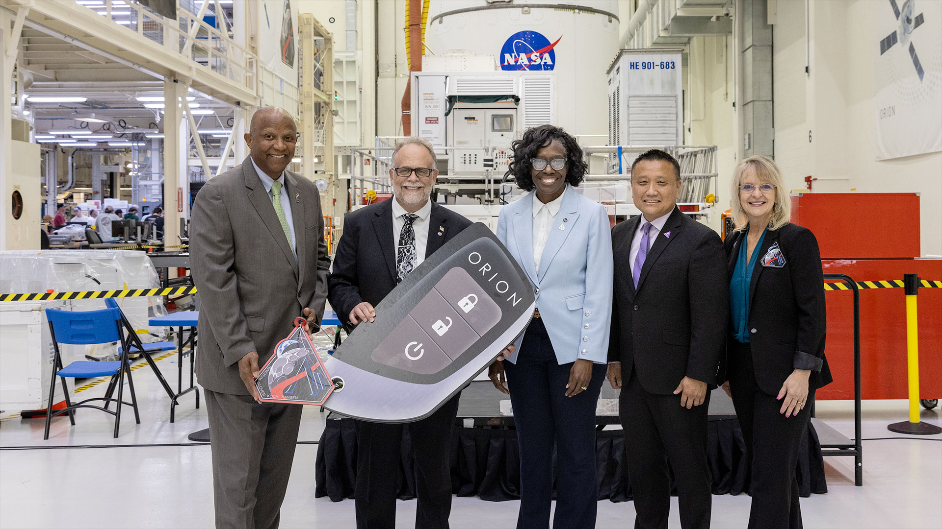 three men and two women pose together standing in front of a spacecraft and holding a ceremonial enlarged key fob.