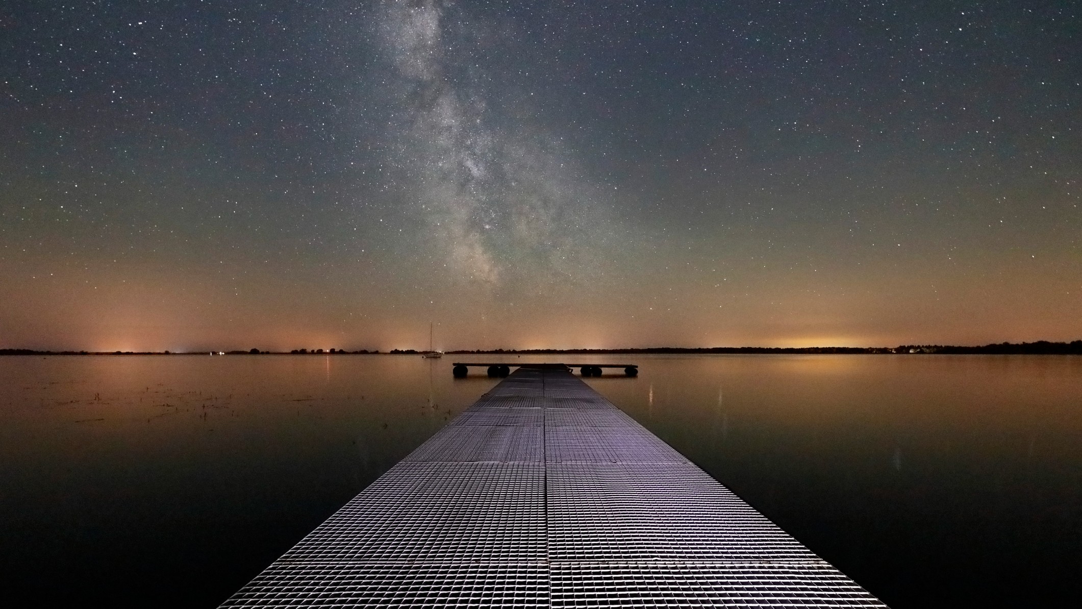 The glowing band of the Milky Way is visible in the sky above a jetty built over a placid lake. Light pollution is seen framing the dark outline of the far shore.