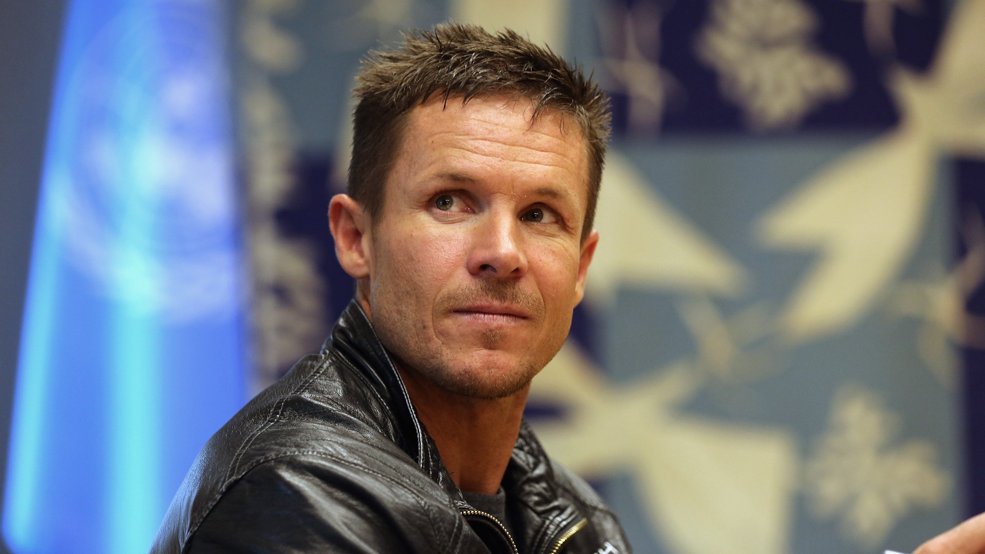 a man with short hair in a leather jacket smiles for a portrait in front of the united nations' flag