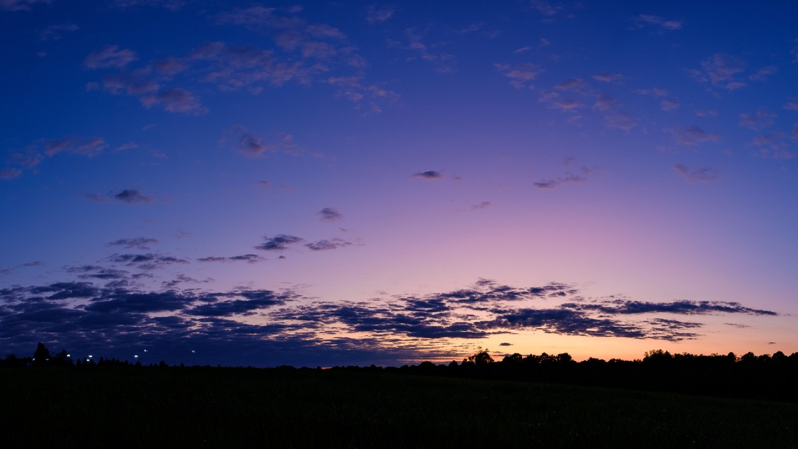An image of the sky in summer as the sun sets below the horizon, giving rise to a variety of hues spanning from orange to deep purple. Clouds can be seen in the sky above the dark silhouette of the horizon.