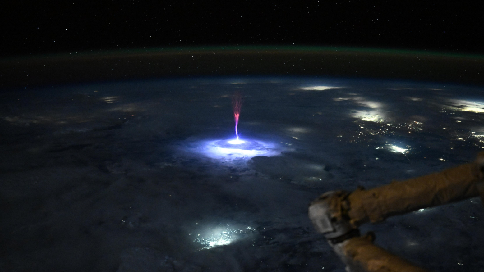 A picture of Earth's surface at night captured from the International Space Station. An electrical phenomenon known as a sprite, featuring a tree-like column and red, branch-like tendrils can be seen reaching into the upper atmosphere above lightning-illuminated clouds.