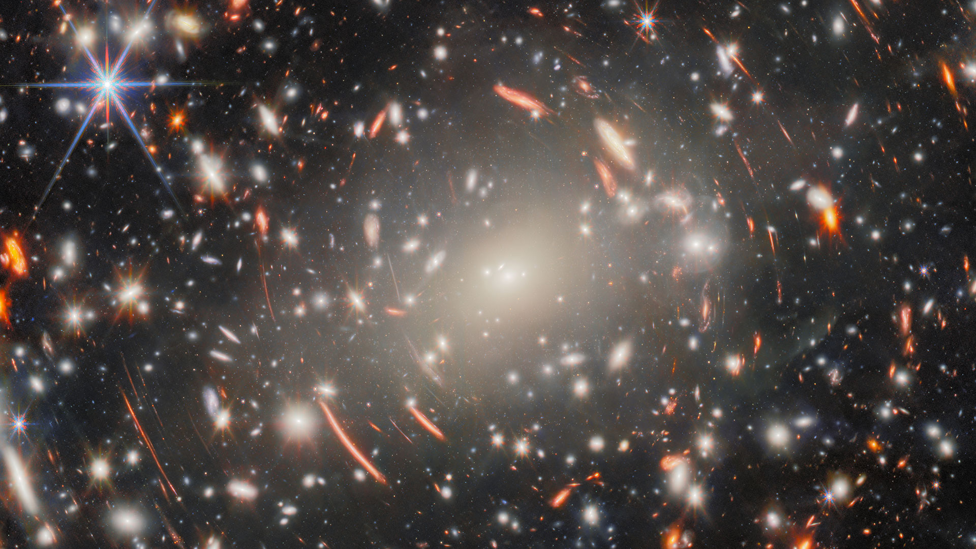 a field of galaxies in space, dominated by an enormous, bright-white elliptical galaxy that is the core of a massive galaxy cluster, surrounded by short, curved, glowing red lines, which are images of distant background galaxies magnified and warped by gravitational lensing.