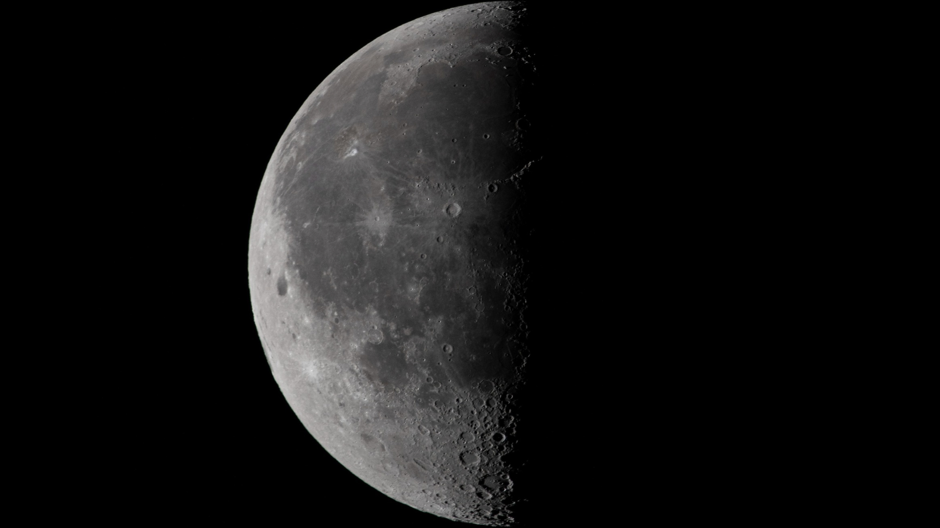 A half-lit moon is pictured against a black sky. The left side of the lunar disk is bathed in sunlight, while the left is wreathed in shadow.