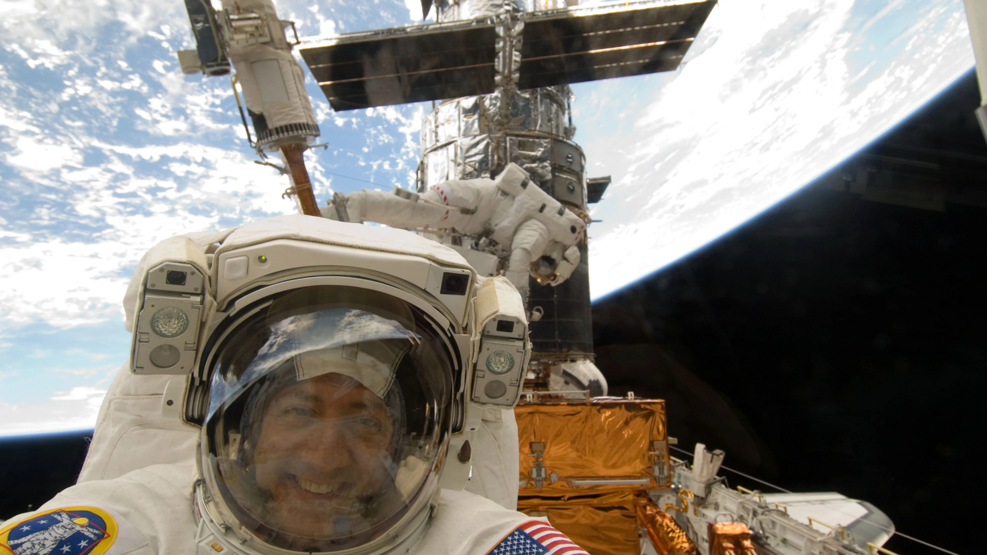 NASA astronaut Mike Massimino smiles for a photo during STS-125 — the fifth and final Hubble Space Telescope servicing mission flown by NASA’s space shuttle fleet — in May 2009. The iconic observatory and Mike Good, Massimino’s spacewalk partner, can be seen in the background.