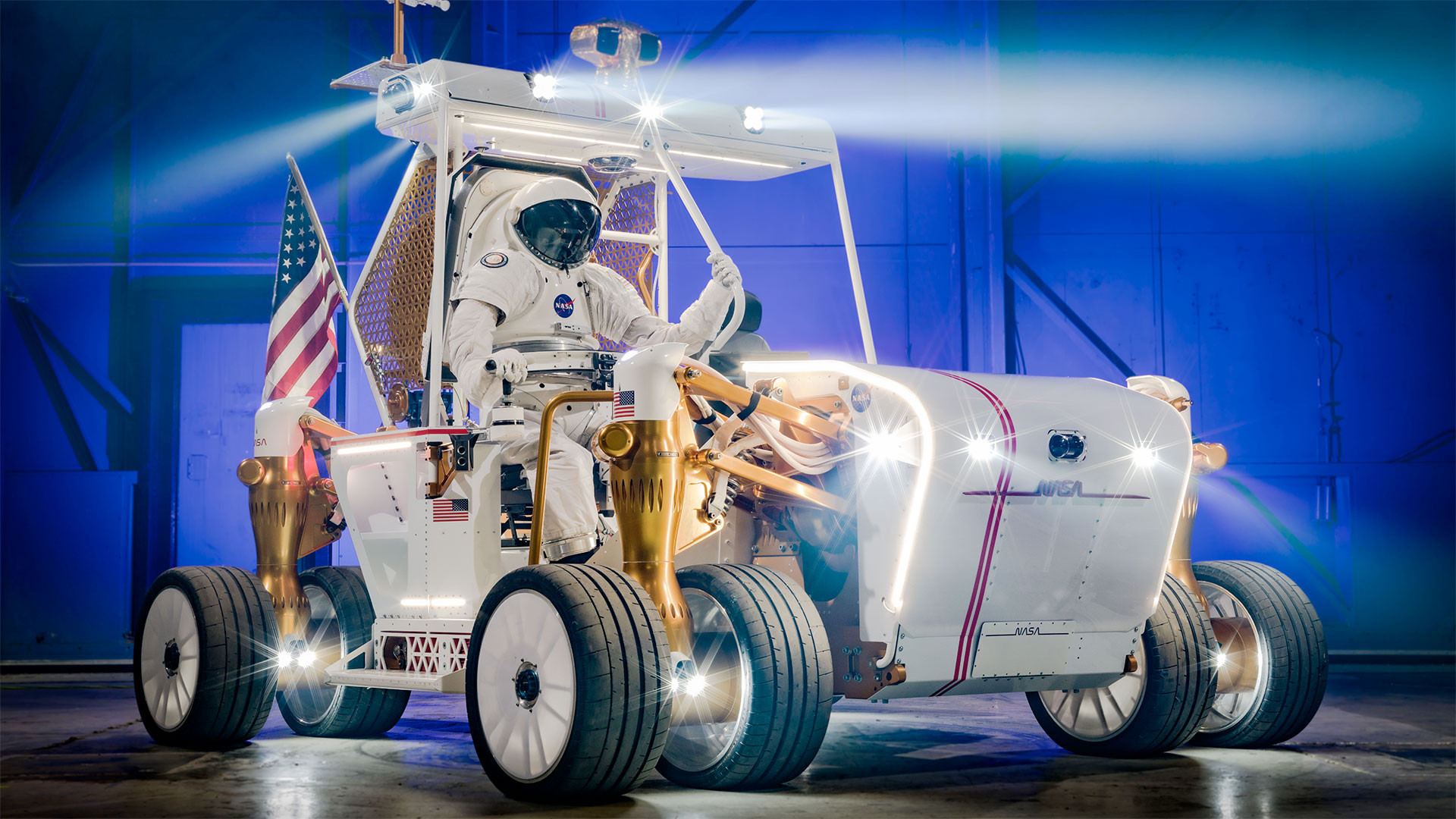 an eight-wheeled moon buggy with a spacesuited astronaut at its controls is seen in a building with its lights turned on.