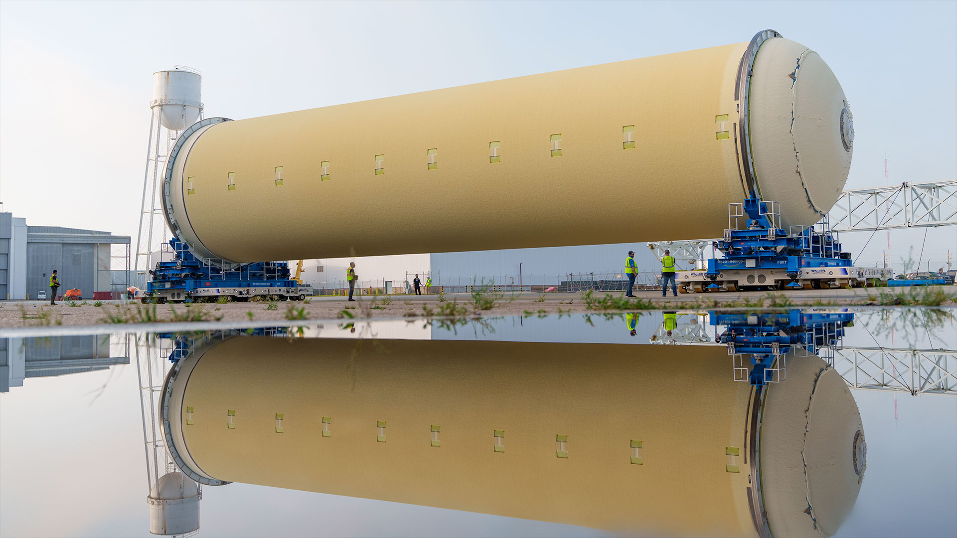 a large, light orange cylindrical tank is moved outside a factory, where a standing body of water catches its reflection