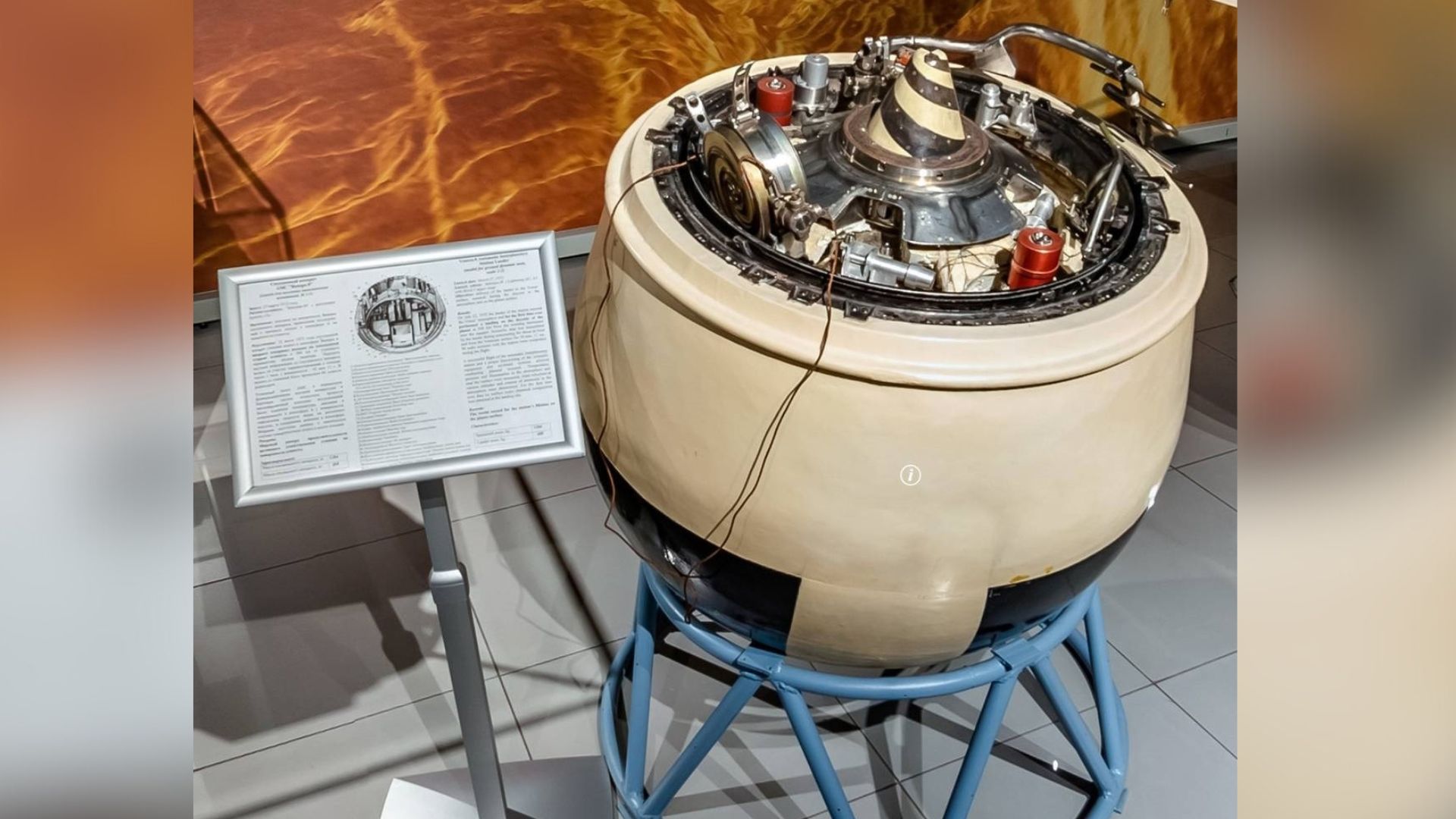 A metal space capsule sits next to a plaque in a museum