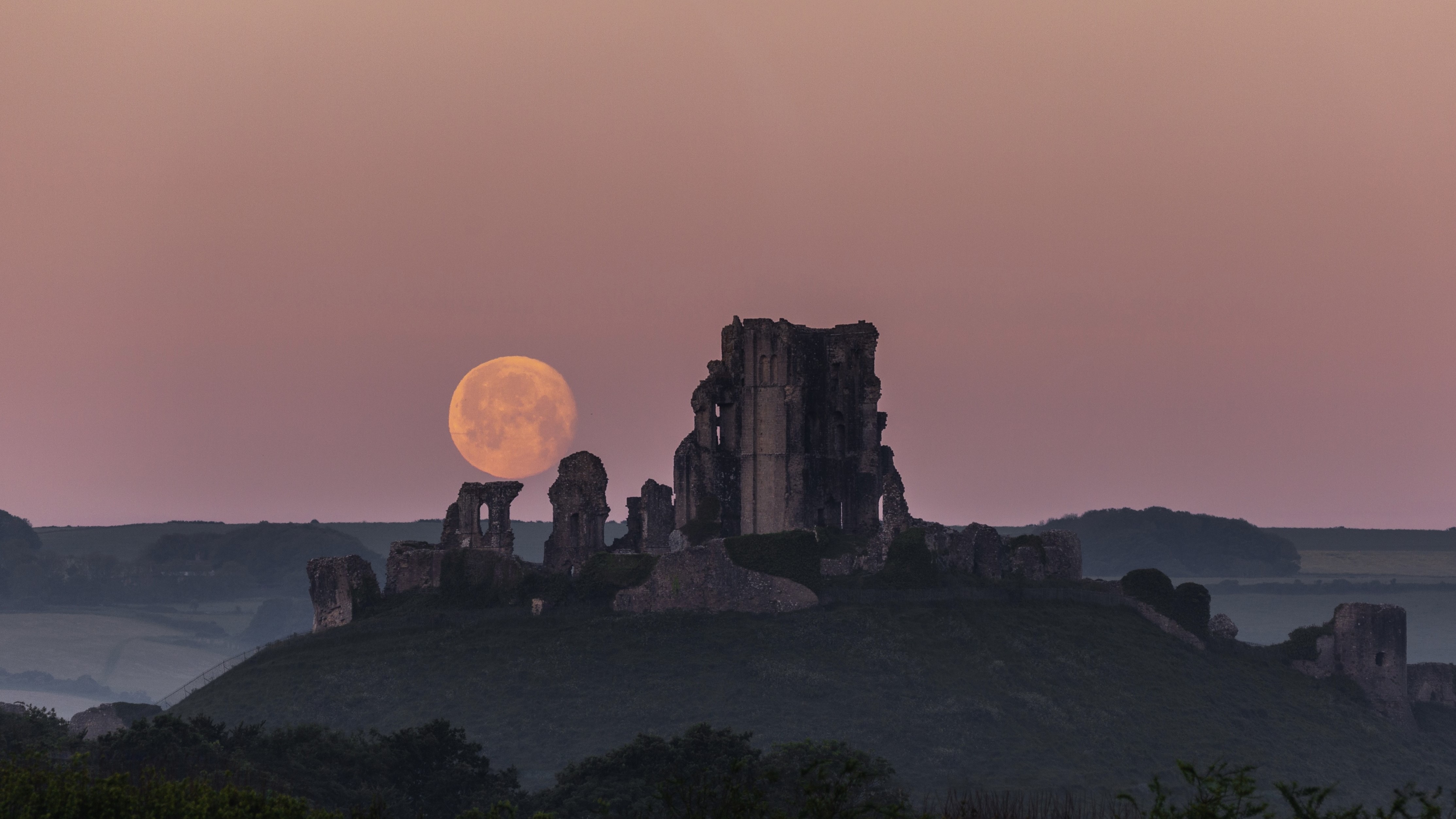 a full moon in a pink hued sky behind a castle ruin.