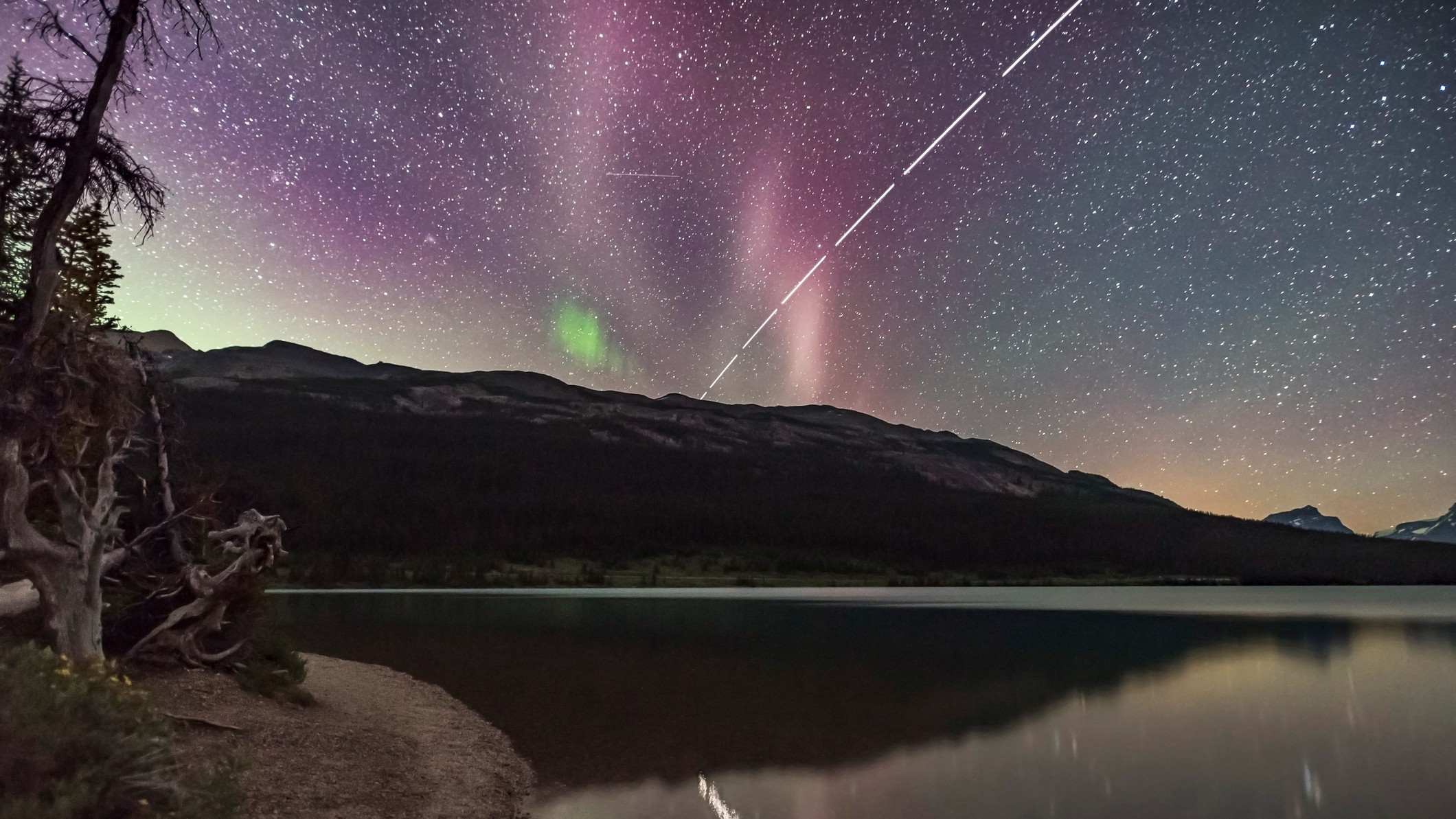 Aurora, stars and satellite streaks can be seen in the sky overlooking a hillside above a lake at night. The bright trail of the International Space Station can also be seen cutting through the sky.